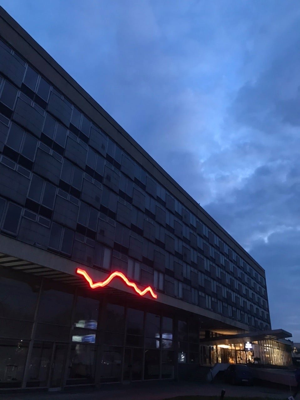 Wielopiętrowy budynek z regularnym układem okien zajmuje kadr ukośnie od lewego dolnego do prawego górnego rogu na tle niebiesko-szarego, zachmurzonego nieba, z czerwonym falistym neonem nad przeszklonym parterem, oświetlonym wejściem po prawej i zaparkowanym samochodem przed wejściem.