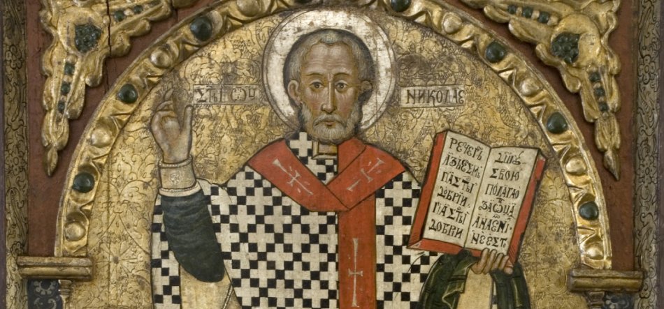 Ikona przedstawiająca postać z aureolą w liturgicznych szatach z czarno-białym kratowanym wzorem i czerwonym ornatem, prawą ręką uniesioną w geście błogosławieństwa, lewą trzymającą otwartą książkę z tekstem w cyrylicy, na złocistym, bogato zdobionym tle z ornamentalną ramą i zielonymi kamieniami.