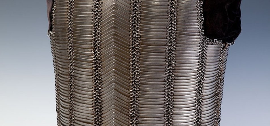 Fragment pancerza z równoległych metalowych listew nachodzących na siebie i połączonych pionowymi ogniwami, ułożonych w wzór w kształcie litery V, z ciemnym materiałem u górnej krawędzi i jasnoszarym tłem.