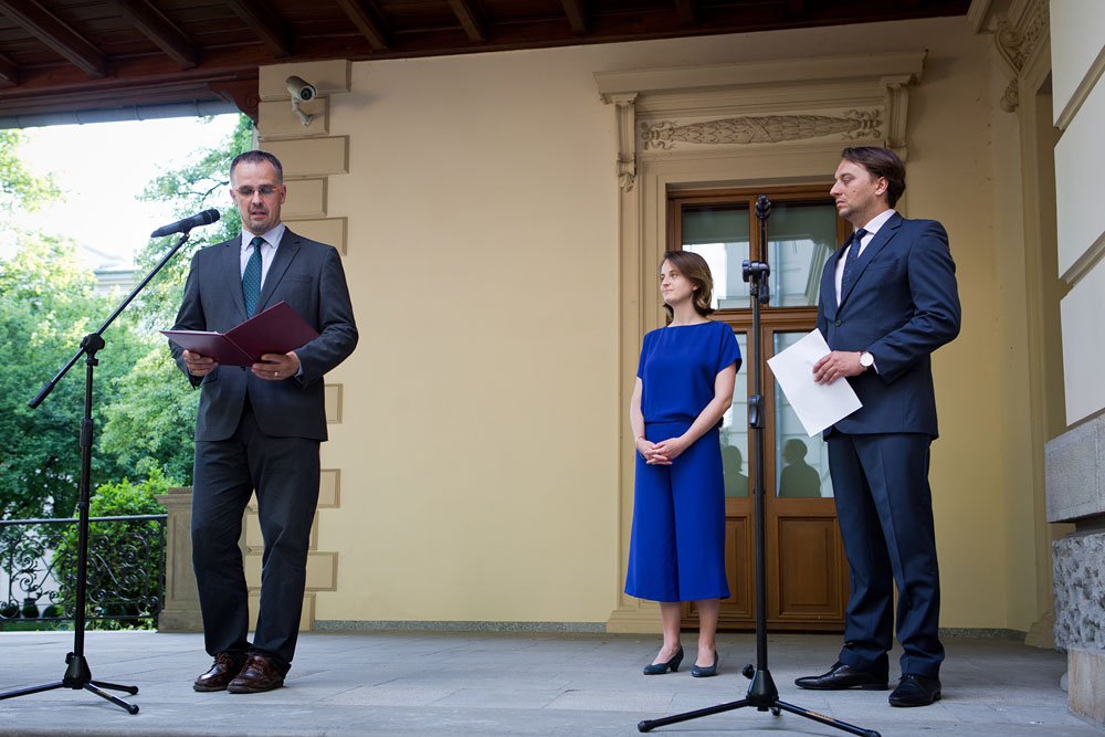 Trzy osoby stoją na zadaszonym tarasie przed drzwiami budynku: po lewej mężczyzna w garniturze przemawia do mikrofonu trzymając otwarty folder, na środku kobieta w niebieskiej sukience stoi ze złożonymi rękami, po prawej mężczyzna w garniturze trzyma kartki przy drugim mikrofonie; w tle beżowa elewacja z ozdobnym portalem, oknem, kamerą i widoczną zielenią oraz metalową balustradą.