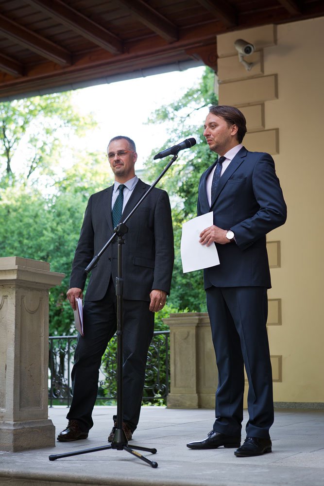 Dwóch mężczyzn w garniturach stoi przy mikrofonie na stojaku na zadaszonym tarasie z kamienną balustradą i zielonymi drzewami w tle, jeden trzyma kartki papieru, na ścianie obok widoczna kamera bezpieczeństwa.