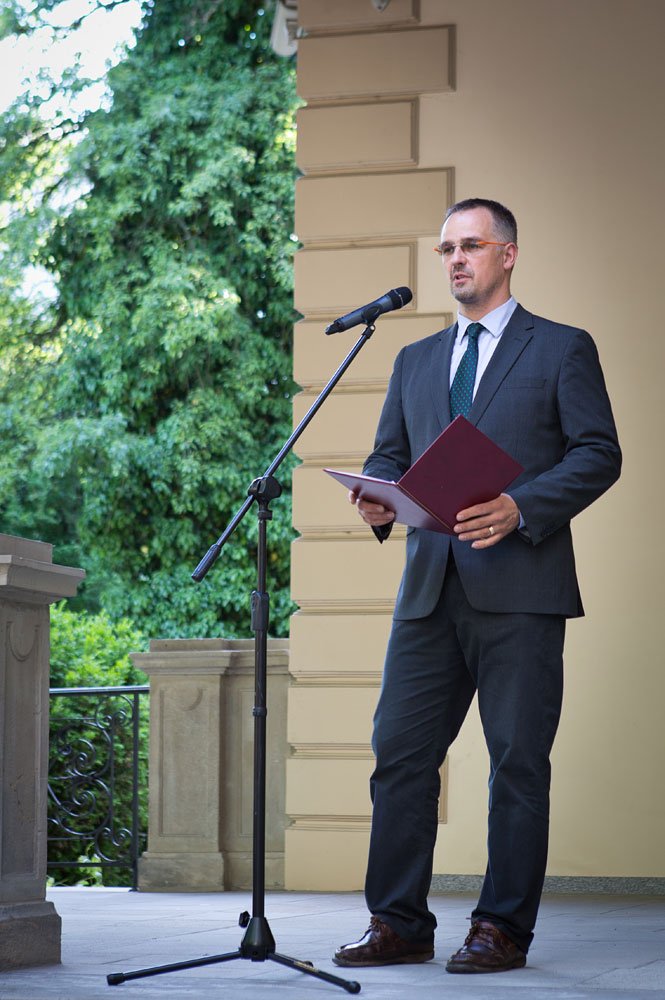 Mężczyzna w ciemnym garniturze, krawacie i okularach stoi przy mikrofonie na statywie, trzymając otwartą bordową teczkę na tle beżowej elewacji budynku, kamiennego balkonika i zielonych drzew.