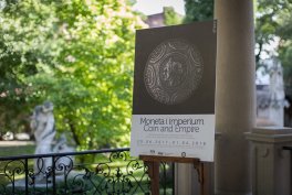 Plansza wystawowa na sztaludze z dużą okrągłą grafiką monety u góry i napisem "Moneta Imperium / Coin and Empire" poniżej, ustawiona na zadaszonym tarasie z ozdobną kutą balustradą, kolumną po prawej i zielonym ogrodem w tle.