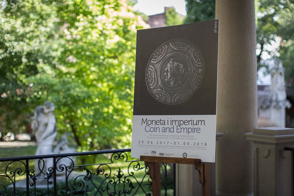 Plakat wystawowy na sztaludze na zadaszonym tarasie, na plakacie duże zdjęcie antycznej monety oraz tekst "Moneta i imperium / Coin and Empire" z datami 29.06.2017–01.04.2018, w tle ozdobne żelazne balustrady, kolumna, rozmyte drzewa i kamienna rzeźba.