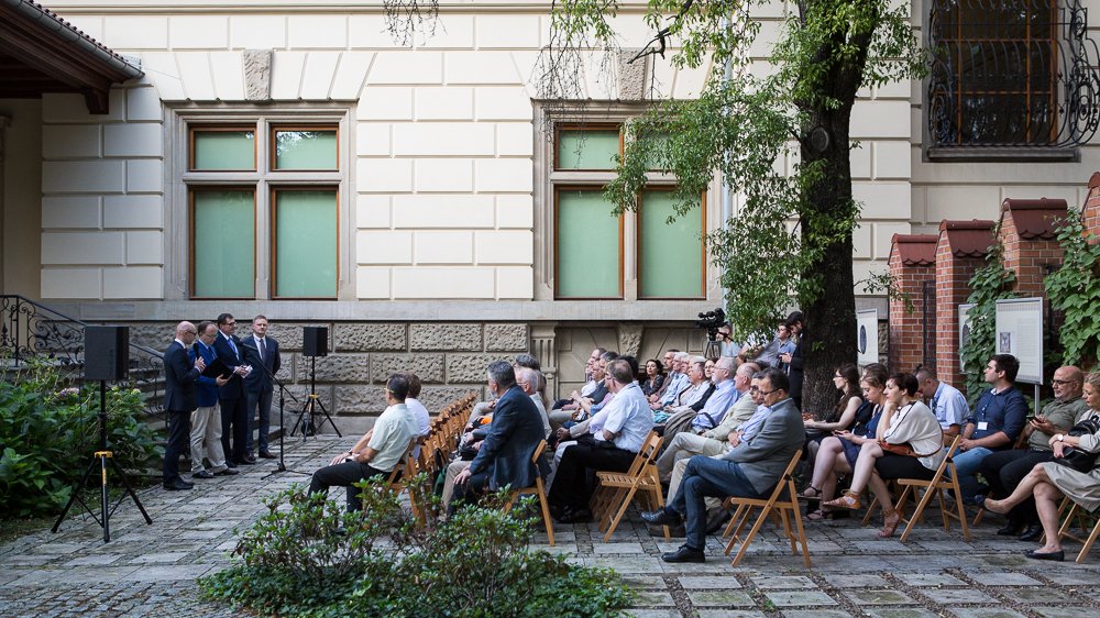 Kamienny dziedziniec z rzędami drewnianych krzeseł, na których siedzi publiczność zwrócona w stronę czterech mężczyzn przemawiających przy mikrofonach po lewej, na tle jasnej elewacji budynku z dwoma dużymi oknami, z drzewem i tablicami wystawowymi po prawej.