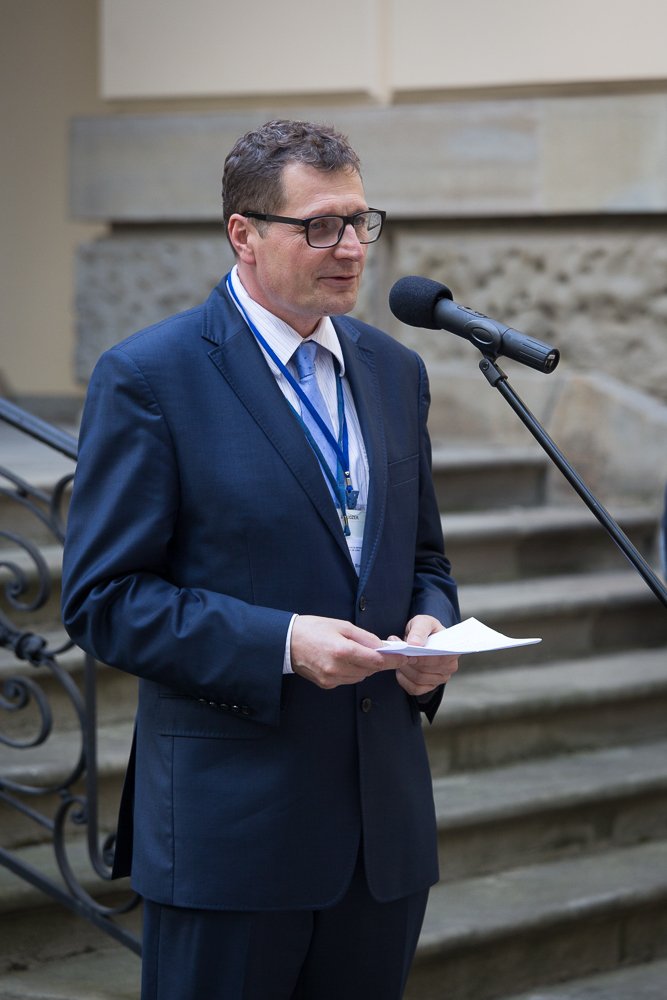 Mężczyzna w granatowym garniturze i okularach z identyfikatorem na niebieskiej smyczy, trzymający kartki i przemawiający do stojącego mikrofonu przed kamiennymi schodami i metalową balustradą.