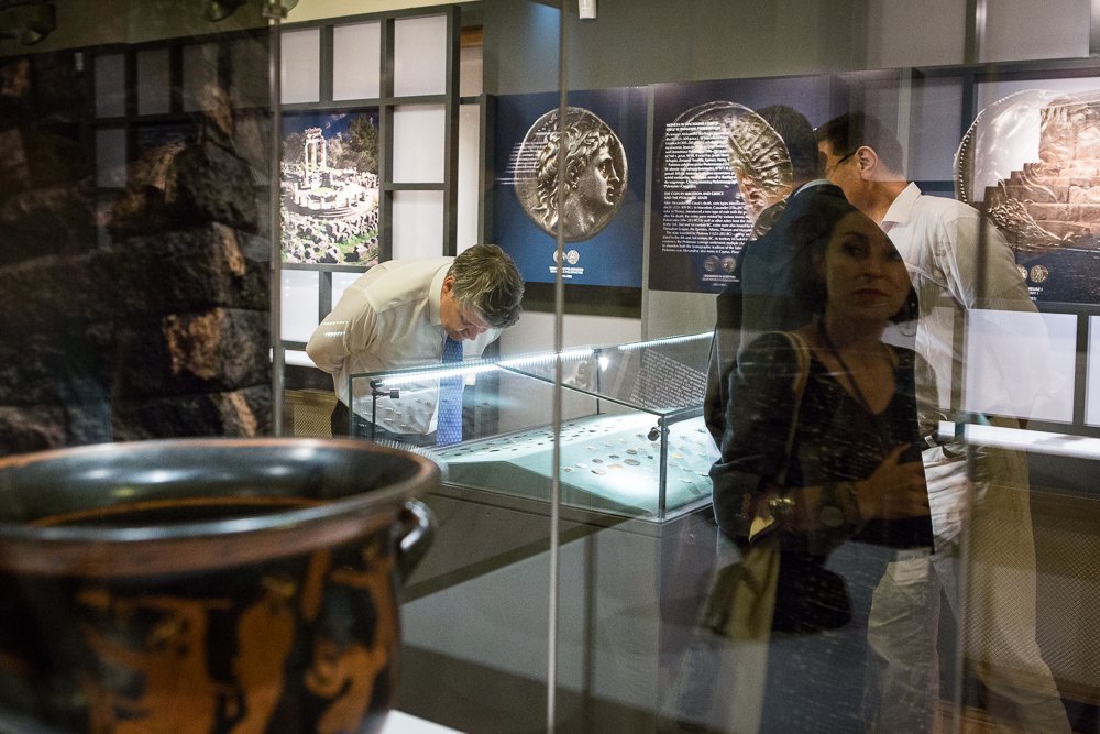 Wnętrze muzeum z przeszkloną gablotą z monetami, mężczyzną pochylonym nad eksponatami, odbiciami innych zwiedzających na szybie, planszami z powiększonymi wizerunkami monet na ścianie i dużym ceramicznym naczyniem na pierwszym planie.