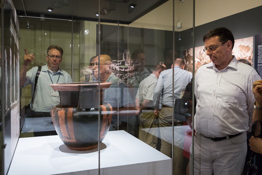 Duży ceramiczny czarno‑czerwony wazon na białym piedestale w szklanej gablocie w muzeum, z kilkoma zwiedzającymi i ich odbiciami w szybach, w tym mężczyzna z identyfikatorem wskazujący w lewo oraz mężczyzna po prawej przyglądający się eksponatowi.