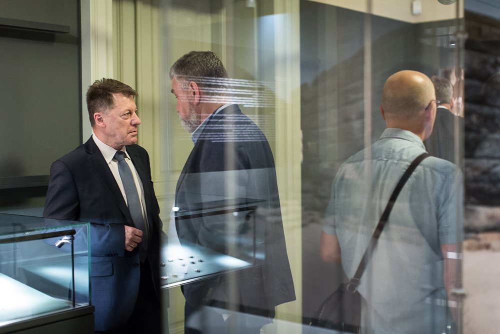 Dwie rozmawiające osoby w garniturach przy oświetlonych szklanych gablotach z drobnymi eksponatami — z lewej mężczyzna w ciemnym garniturze i krawacie, naprzeciw niego mężczyzna z brodą w marynarce, po prawej odwrócony plecami łysy mężczyzna w jasnej koszuli z torbą na ramieniu, całość sfotografowana przez refleksy szyb.