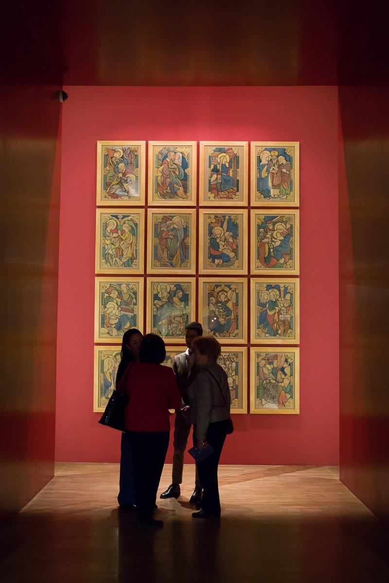 Cztery osoby w półmroku stoją na drewnianej podłodze przed czerwoną ścianą galerii, na której w złotych ramkach ułożono 16 kolorowych figuratywnych obrazów w czterech rzędach i czterech kolumnach, a scena otoczona jest ciemnymi, brązowymi ścianami bocznymi.