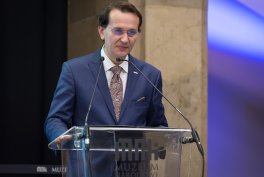 Mężczyzna w granatowym garniturze, wzorzystym krawacie, poszetce i okularach stoi za przezroczystą mównicą z mikrofonem, z rękami na jej krawędzi, na tle kamiennej kolumny i rozmytego niebiesko-fioletowego oświetlenia.