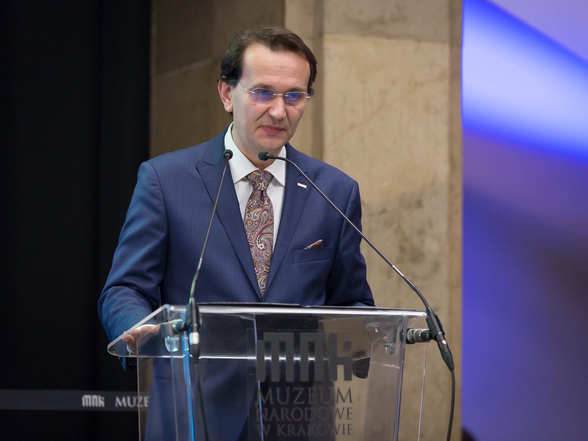 Mężczyzna w granatowym garniturze, białej koszuli, paisley krawacie i okularach stoi za przezroczystą mównicą z dwoma mikrofonami, na przedniej ściance mównicy widoczny napis MUZEUM NARODOWE W KRAKOWIE, w tle kamienna ściana i niebieskie oświetlenie.