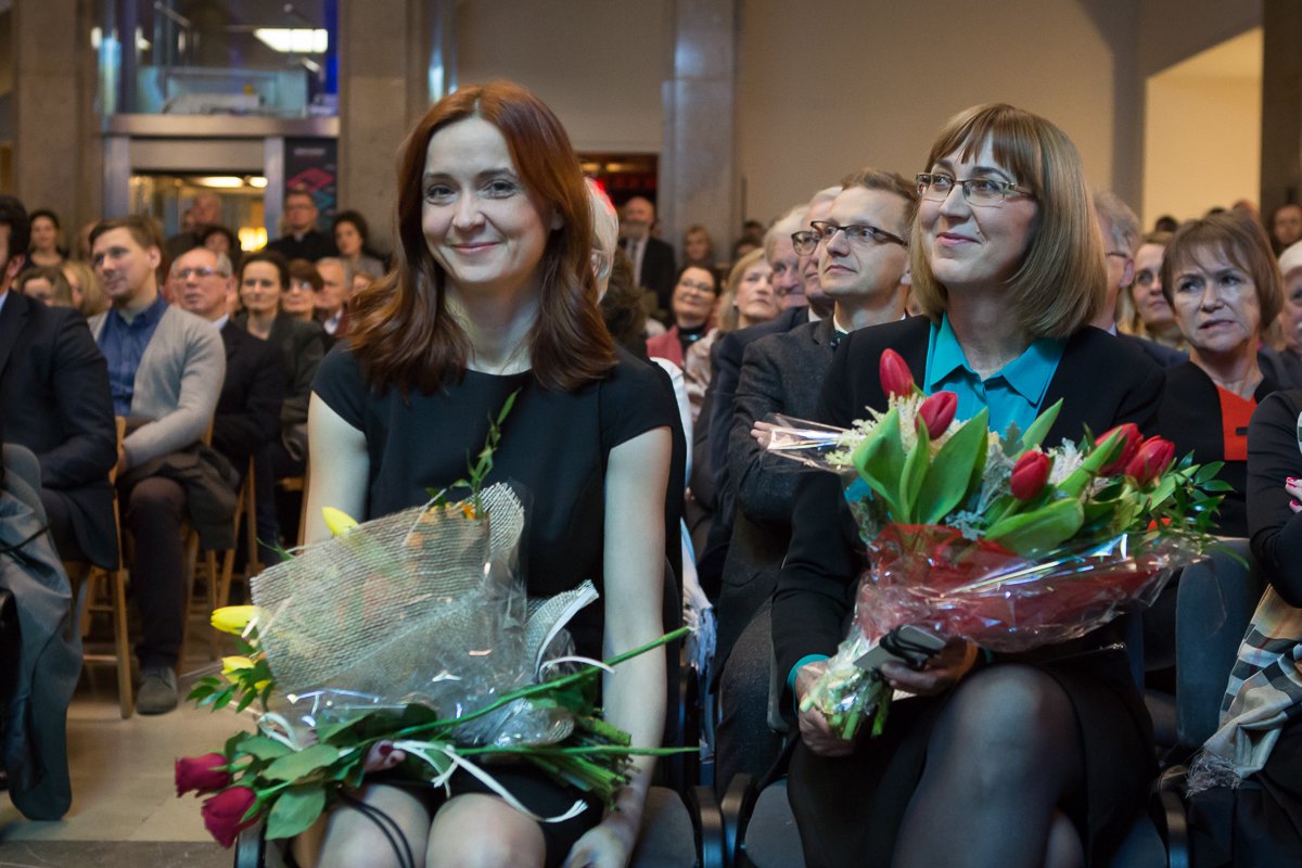 Dwie kobiety siedzą w pierwszym rzędzie sali z liczną widownią w tle: po lewej kobieta o rudych włosach w czarnej sukience trzyma bukiet owinięty siatką, po prawej kobieta w okularach i turkusowej bluzce trzyma owinięty w folię bukiet czerwonych tulipanów oraz telefon.
