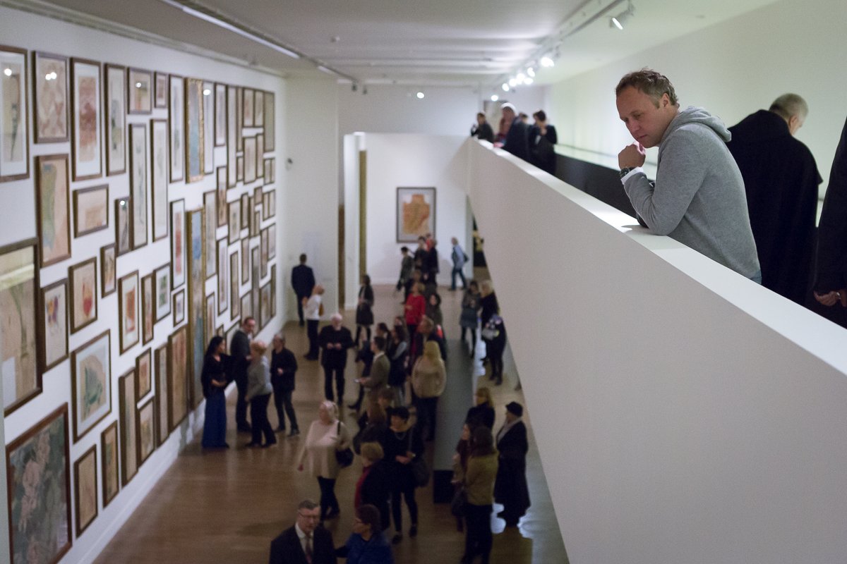 Wnętrze galerii sztuki z długą białą balustradą po prawej, mężczyzną w szarej bluzie opartym o jej krawędź, poniżej kilkudziesięcioma odwiedzającymi spacerującymi po drewnianej podłodze i oglądającymi ścianę po lewej wypełnioną wieloma oprawionymi obrazami ułożonymi w gęstą kompozycję.