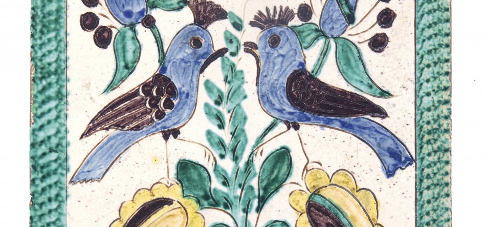 Ceramiczna płytka z dekoracyjnym motywem dwóch niebieskich ptaków zwróconych ku sobie, z ciemnymi detalami piór i dziobów, siedzących na zielonych łodygach między zielonymi liśćmi i żółtymi kwiatami na kremowym tle, otoczona zielonym fakturowanym obramowaniem.