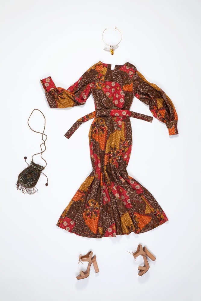 Flat lay: długa sukienka maxi w brązowo-czerwono-pomarańczowy wzór patchwork z bufiastymi rękawami i paskiem ułożona na białym tle, powyżej złoty naszyjnik z zawieszką, po lewej mała koralikowa torebka na sznurku, na dole beżowe sandały na słupku.