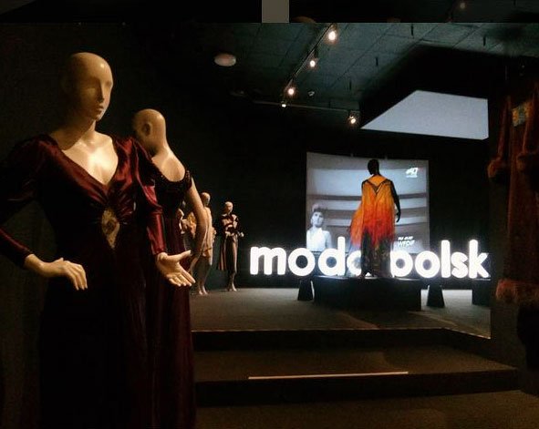 W ciemnej sali wystawowej po lewej kilka manekinów w długich sukienkach, na scenie podświetlony napis "moda polsk", na podwyższeniu sylwetka w pomarańczowo‑żółtym płaszczu przed ekranem z czarno‑białą projekcją.