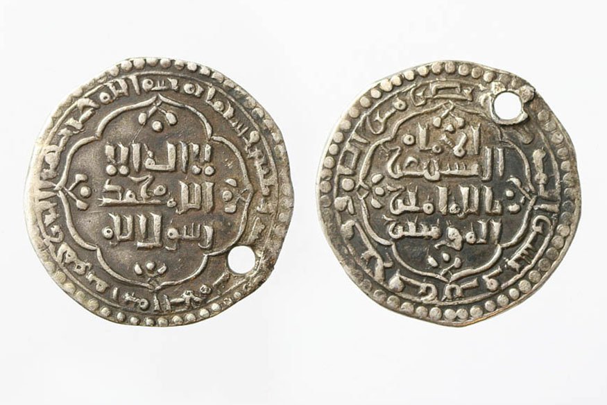 Zdjęcie przedstawia obie strony metalowej, srebrnoszarej monety ułożone obok siebie na białym tle; każda strona ma falistą ozdobną ramkę z gęstym arabskim pismem i kropkowanym obrzeżem, przy czym na obu krążkach widoczny jest otwór przebity blisko krawędzi.