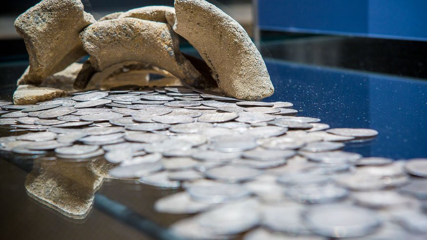 Liczne metalowe monety rozsypane na błyszczącej szklanej powierzchni z widocznym odbiciem; w tle łukowato ułożone jasne fragmenty ceramiki i rozmyte niebieskie tło.