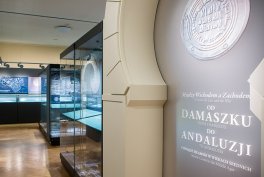 Wnętrze sali wystawowej z szeregiem podświetlonych gablot po lewej, drewnianą podłogą i jasnym łukiem ściennym po prawej, na którym widoczna okrągła pieczęć oraz napis "OD DAMASZKU DO ANDALUZJI".