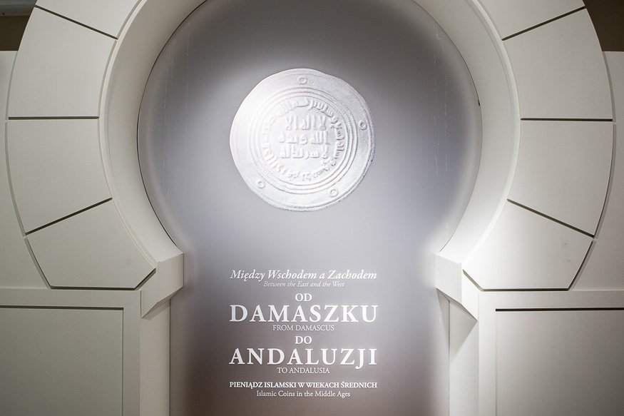 Wnęka wystawowa z białą arkadową ramą, wewnątrz szara płaszczyzna z dużym odciskiem monety u góry oraz białym tekstem po polsku i angielsku informującym: „Między Wschodem a Zachodem / Od Damaszku do Andaluzji” i dopiskiem o pieniądzu islamskim w wiekach średnich.