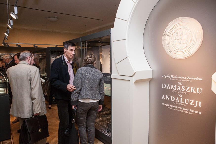 Wnętrze muzeum z kilkoma zwiedzającymi stojącymi przy szklanych gablotach z eksponatami, sufitowe reflektory, drewniana podłoga i po prawej łukowa oprawa panelu wystawy z dużym wizerunkiem monety i napisem „OD DAMASZKU DO ANDALUZJI”.