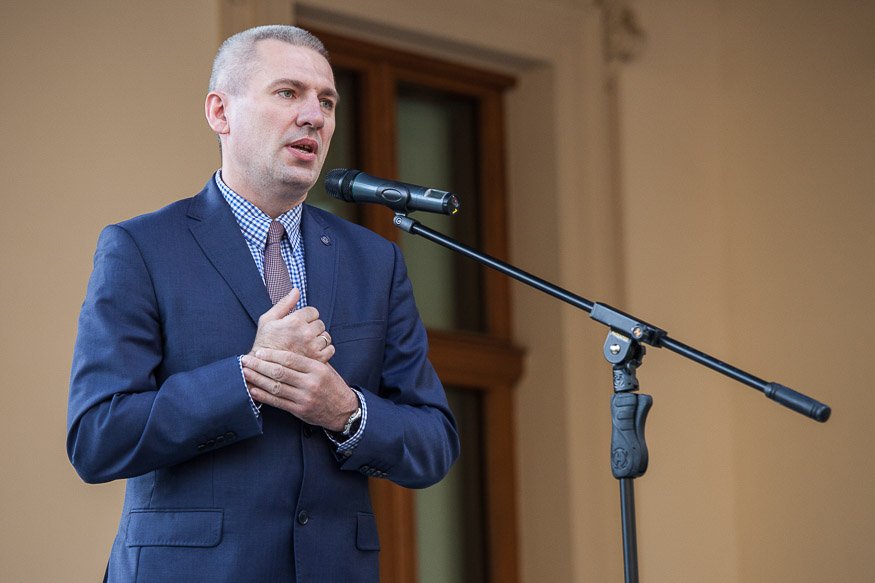 Mężczyzna w granatowym garniturze, kraciastej koszuli i brązowym krawacie stoi przy mikrofonie na wysięgniku, mając splecione dłonie przy piersi, na tle beżowej ściany i drewnianego okna.