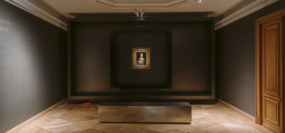 Wnętrze eleganckiej galerii o ciemnych ścianach z centralnie wyeksponowanym małym portretem w ozdobnej złotej ramie w głębi, przed którym stoi niski prostokątny błyszczący podest na jasnym parkiecie; po lewej widoczny fragment ściany z tekstem informacyjnym, po prawej drewniane drzwi i ozdobny sufit.