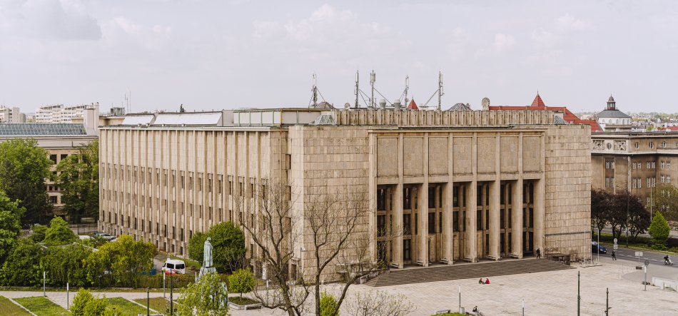 Duży prostokątny budynek z jasnego kamienia z wysokimi pionowymi filarami i szerokimi schodami wejściowymi, na pierwszym planie otwarty plac z kilkoma osobami, drzewami, posągiem i zaparkowanymi samochodami, a w tle dachy miasta z antenami.