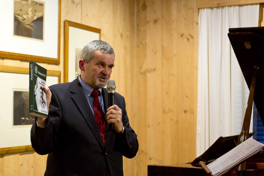 Mężczyzna z siwymi włosami w ciemnym garniturze i czerwonym krawacie trzyma mikrofon przy ustach i zieloną książeczkę z czarno-białym zdjęciem na okładce, stoi przed drewnianą ścianą z oprawionymi obrazami, obok widoczny fortepian z nutami i białe zasłony.