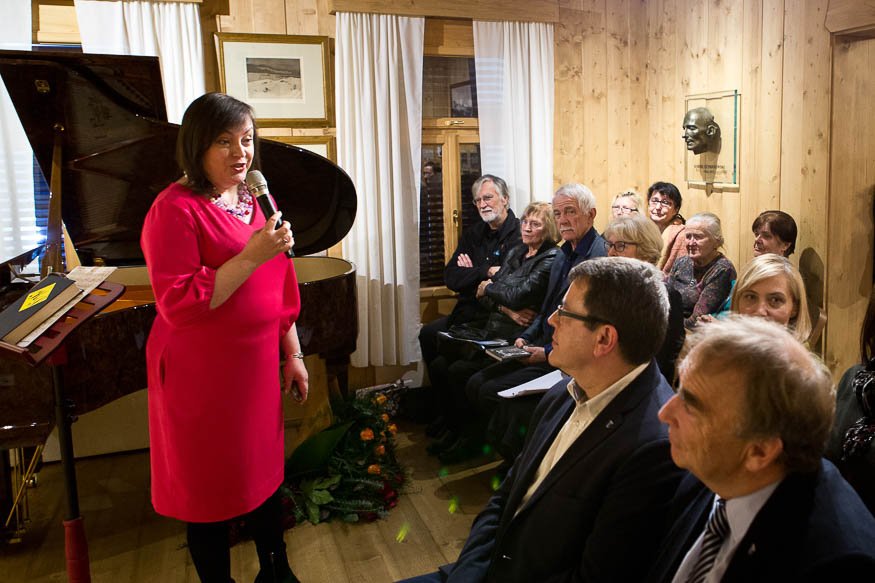 Kobieta w różowej sukience stoi z mikrofonem przy otwartym fortepianie i przemawia do siedzącej publiczności w małym, drewnianym pomieszczeniu z białymi zasłonami, obrazami na ścianie, popiersiem i kwiatowym wieńcem na podłodze.
