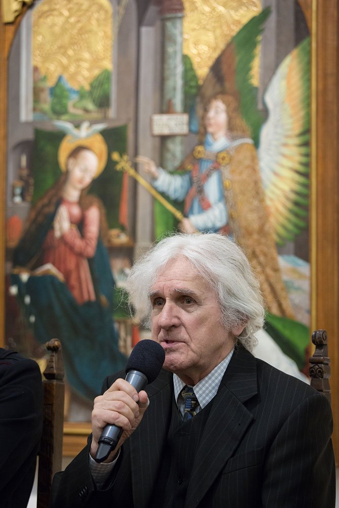 Starszy mężczyzna o siwych włosach trzyma mikrofon przy ustach, ubrany w ciemny garnitur i krawat, siedzi na krześle na tle dużego religijnego obrazu przedstawiającego Marię z aureolą i anioła z rozpostartymi skrzydłami na złotym tle.