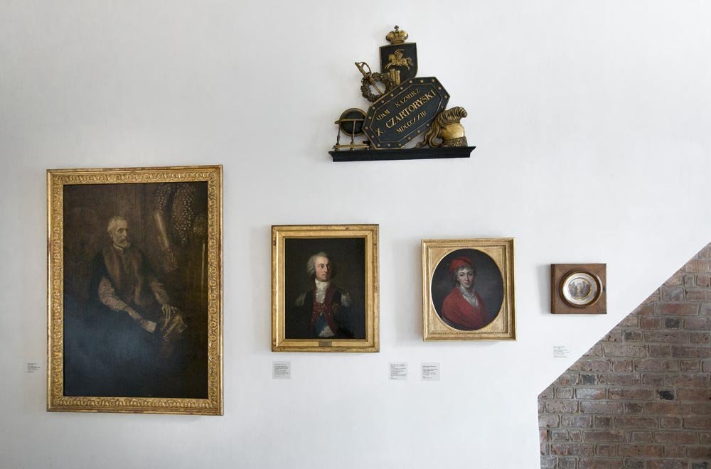 Ściana galerii z białym tłem i fragmentem czerwonej ceglanej ściany po prawej, na której wiszą cztery oprawione obrazy — duży złocony portret siedzącej postaci po lewej; dwa mniejsze złocone portrety pośrodku (prostokątny portret mężczyzny w stroju oficerskim oraz okrągły portret kobiety w czerwonym nakryciu) i mała okrągła praca w drewnianej ramie po prawej; nad środkowymi obrazami zawieszona czarno‑złota herbowa tablica z koroną i ozdobnymi elementami, pod obrazami widoczne etykiety muzealne.