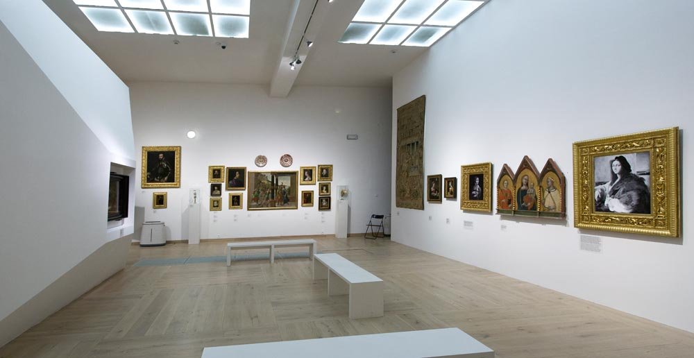 Wnętrze jasnej galerii sztuki z białymi ścianami i jasną drewnianą podłogą, kilkoma białymi ławkami pośrodku, a na ścianach z prawej i tylnej strony ułożone w ramy obrazy (w tym tryptyk), wiszący arras oraz drobne rzeźby na postumentach pod sufitowymi świetlikami.
