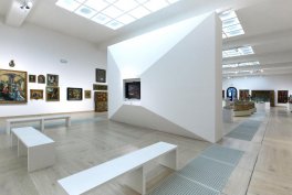 Wnętrze jasnej galerii sztuki z białą geometryczną ścianką z ukośnym wcięciem, białymi ławkami na pierwszym planie, obrazami wiszącymi po lewej, gablotami i rzeźbami w tle oraz przeszklonymi świetlikami w suficie.