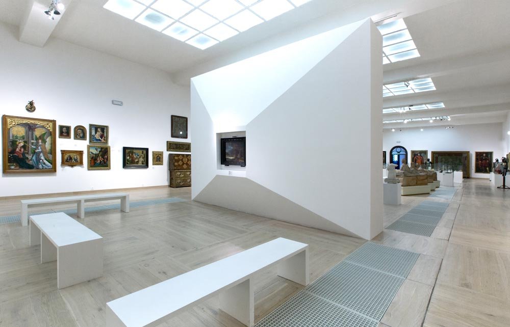 Wnętrze jasnej galerii sztuki z białymi ścianami i jasną drewnianą podłogą, dominującą dużą białą geometryczną ścianką z wcięciem na obraz, kilkoma białymi ławkami na pierwszym planie, obrazami wiszącymi po lewej, rzeźbami na postumentach po prawej, metalowymi kratkami w podłodze, świetlikami w suficie i kilkoma osobami w tle.