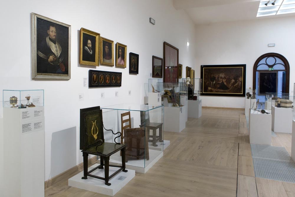 Wnętrze jasnej galerii muzealnej z białymi ścianami i drewnianą podłogą, po lewej ścianie kilka oprawionych portretów, przed nimi krzesła i drobne przedmioty w szklanych gablotach ustawione na białych postumentach, po prawej dalsze eksponaty i duże płótno na końcu sali oraz łukowe przejście i świetlik w suficie.