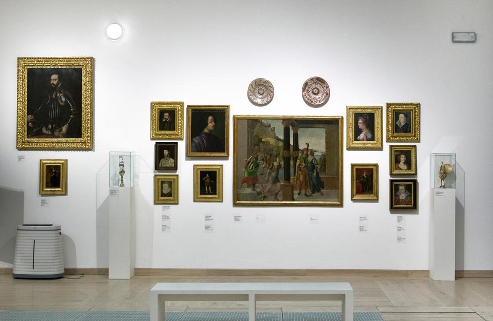 Wnętrze galerii z białą ścianą, na której w symetrycznym układzie zawieszono kilkanaście obrazów w ozdobnych złotych ramach i dwie dekoracyjne płytki nad dużym centralnym płótnem, po bokach dwie przeszklone gabloty z eksponatami oraz niska biała ławka na pierwszym planie.