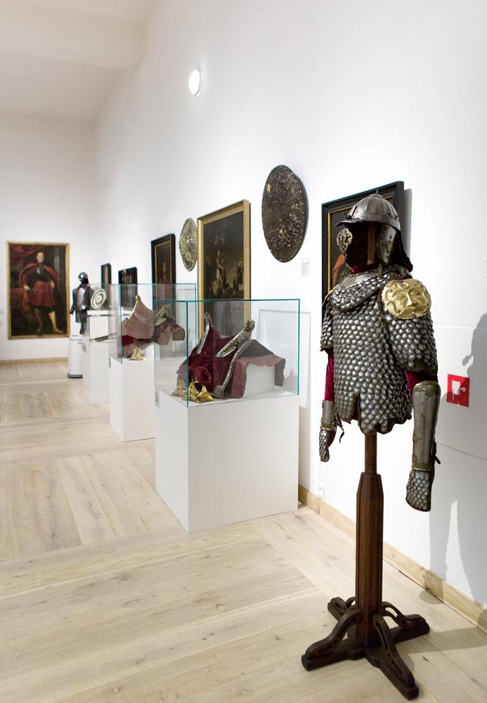Wnętrze galerii muzealnej z jasną drewnianą podłogą i białymi ścianami, po prawej stojak z hełmem i kolczugą z blaszek na drewnianej podstawie, w centrum rząd białych postumentów z przeszklonymi gablotami zawierającymi historyczne hełmy i czerwone tkaniny, a na ścianie po lewej wiszą obrazy i okrągłe tarcze.