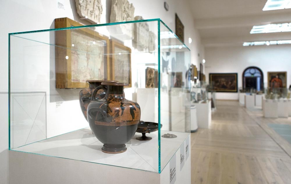 Wnętrze muzealnej sali z pierwszego planu pokazującą szklaną gablotę na białym postumencie zawierającą dwie ceramiczne amfory o czarno‑pomarańczowych dekoracjach i małe naczynie, w tle białe ściany z obrazami i innymi eksponatami oraz drewniana podłoga.