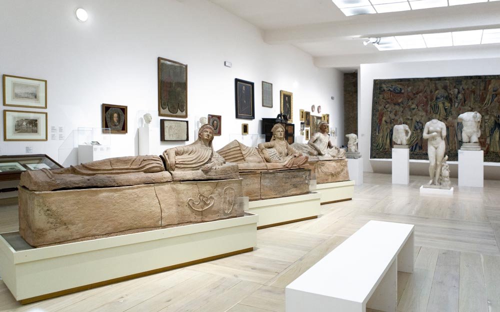 Wnętrze jasnej galerii muzealnej z drewnianą podłogą i świetlikami w suficie; na pierwszym planie trzy rzeźbione leżące sarkofagi na niskich białych postumentach, po prawej kamienne torsy i popiersia na cokole przed dużym, kolorowym gobelinem na tylnej ścianie, po lewej ściana z wiszącymi obrazami i grafikami oraz biała ławka na środku.