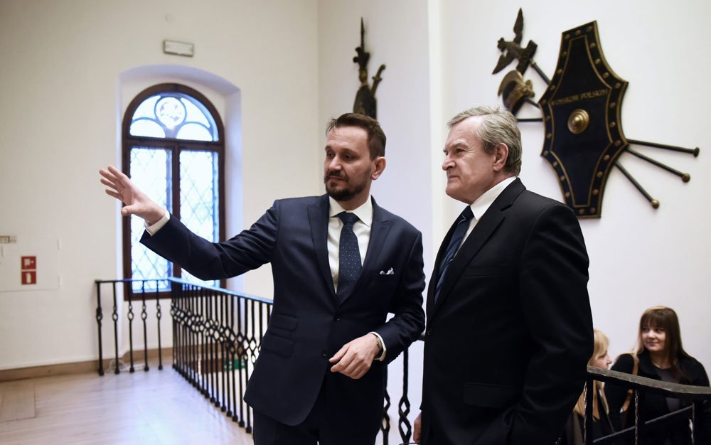 Dwaj mężczyźni w garniturach stoją we wnętrzu z ozdobnymi tarczami na ścianie, jeden gestykuluje w lewo, w tle arkadowe okno z kratą i metalowa balustrada, po prawej częściowo widoczne dwie siedzące kobiety.