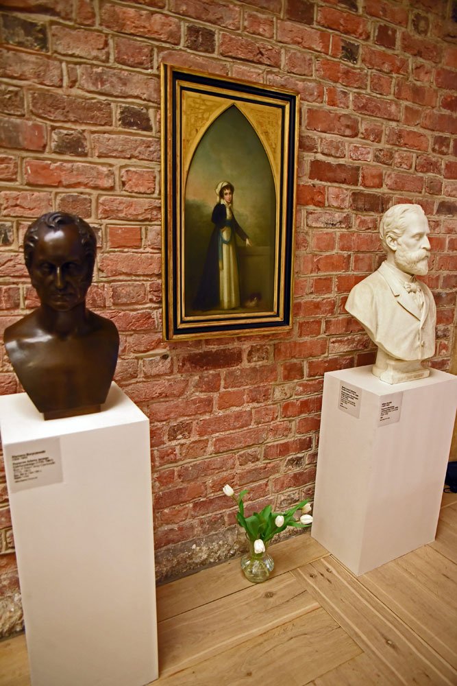 Wnętrze wystawy z ceglaną ścianą: centralnie zawieszony w złotej, ostrołukowej ramie portret kobiety w historycznej sukni, po bokach na białych postumentach dwa popiersia (ciemne, brązowe po lewej i jasne, marmurowe po prawej), na drewnianej podłodze mały szklany wazon z białymi tulipanami oraz widoczne tabliczki informacyjne.