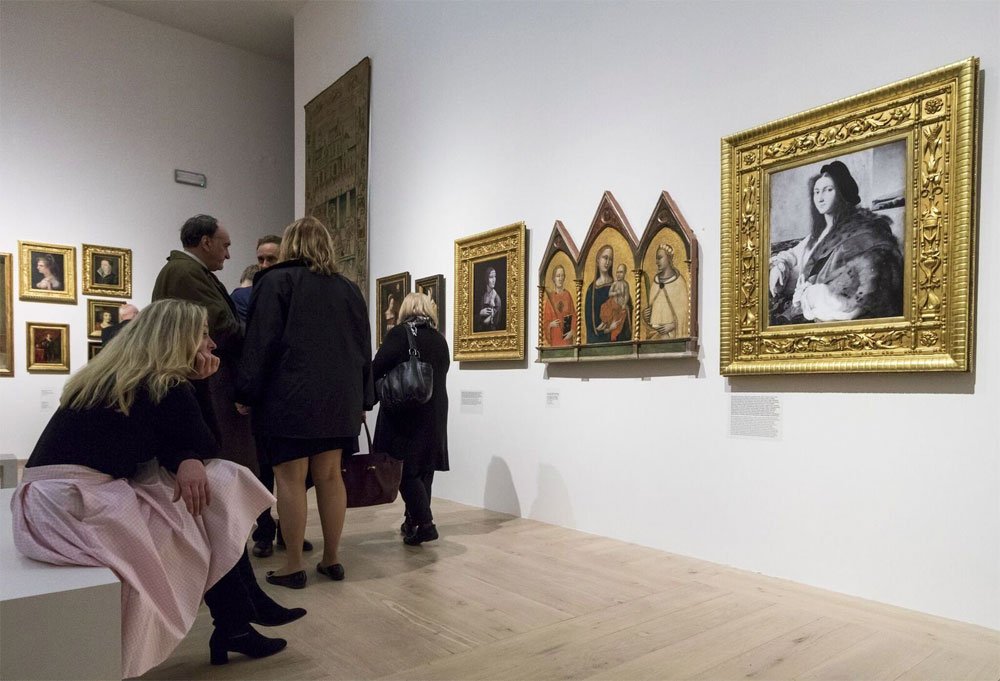 Wnętrze galerii sztuki z jasną drewnianą podłogą, gdzie kilka osób stoi i ogląda oprawione obrazy na białej ścianie; po lewej jedna osoba siedzi na białej ławce, po prawej widoczny duży złocony portret i trójdzielny tryptyk z postaciami.