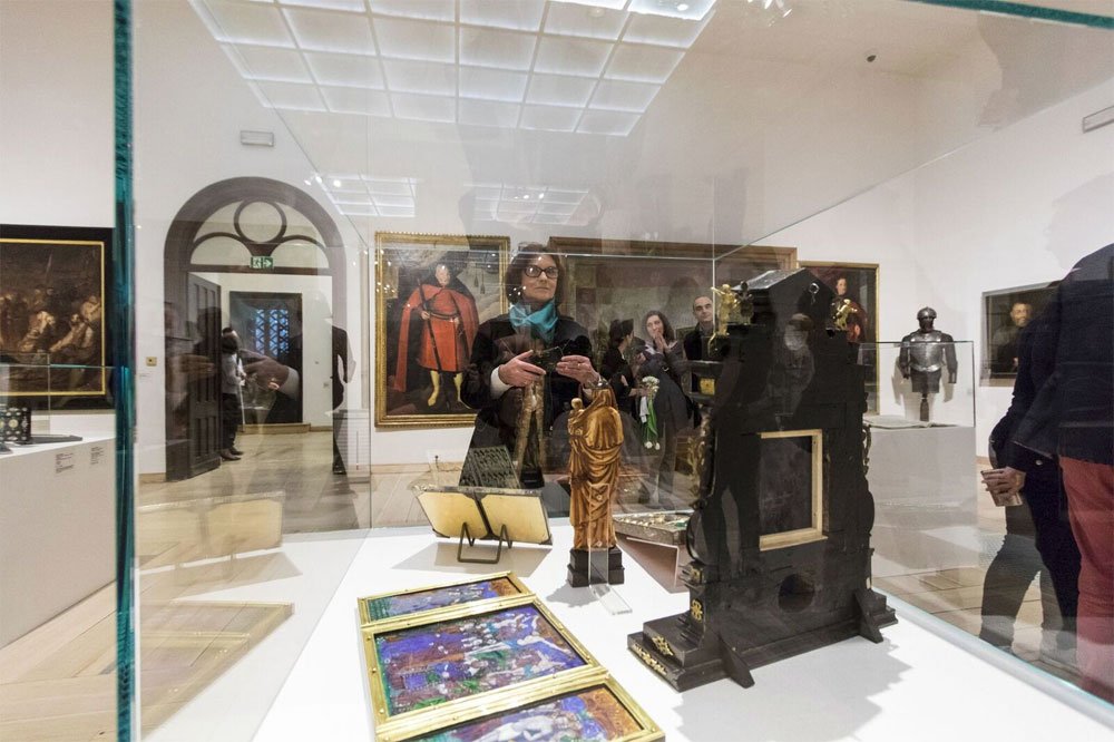 Wnętrze galerii muzealnej widziane przez szklaną gablotę zawierającą małe rzeźby — pozłacaną figurkę i czarną ozdobną konstrukcję — oraz trzy drobne, oprawione obrazy na pierwszym planie; na ścianach w tle portrety i zbroja, a w odbiciu szyby widać kilka osób zwiedzających, w tym osobę w okularach trzymającą telefon.