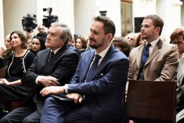 Kilka osób siedzących w rzędach podczas wydarzenia — w pierwszym planie trzech mężczyzn w formalnych strojach (mężczyzna w granatowym garniturze pośrodku, mężczyzna w ciemnym garniturze po lewej i mężczyzna w beżowym płaszczu po prawej), za nimi widoczna publiczność i operatorzy kamer.