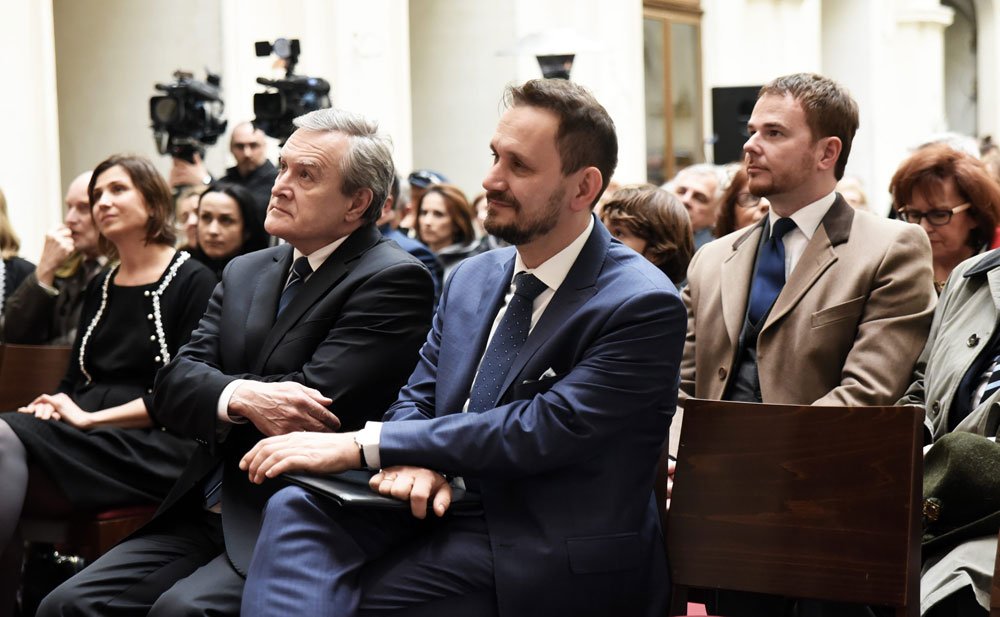 Grupa osób siedzących w rzędach w jasnym pomieszczeniu — na pierwszym planie trzech mężczyzn w formalnych ubraniach (pośrodku w niebieskim garniturze i kropkowanym krawacie, po lewej starszy mężczyzna w ciemnym garniturze, po prawej młodszy w beżowym płaszczu), w tle kobiety i kamery filmowe.