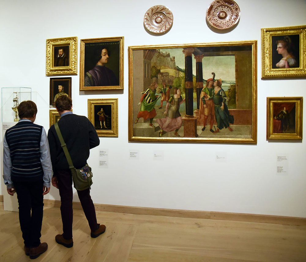 Dwóch zwiedzających stojących tyłem przed białą ścianą galerii z kilkoma złoconymi ramami, dużym obrazem sceny figuralnej w centrum, dwoma ozdobnymi talerzami nad nim, przeszkloną gablotą po lewej, etykietami pod obrazami i jasną drewnianą podłogą.