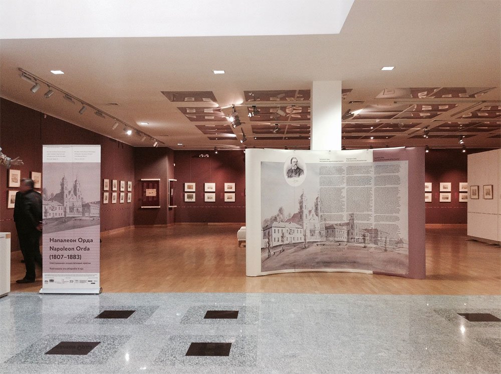 Wnętrze galerii z drewnianą podłogą i połyskującą przednią częścią z szarych płytek, centralnie ustawioną zakrzywioną planszą z dużym rysunkiem budynków i tekstem, po lewej stojak wystawowy z widocznym napisem Napoleon Orda (1807–1883), bordowe ściany w tle z wieloma oprawionymi grafikami, białe słupy, sufit z reflektorami oraz jedna zamazana postać po lewej stronie