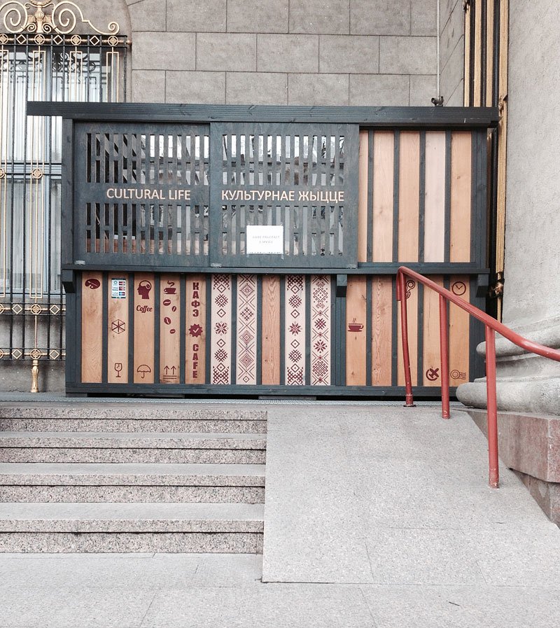 Przed szarą kamienną ścianą stoi drewniana zabudowa z górnym ażurowym panelem z napisem CULTURAL LIFE i cyrylicą, poniżej pionowe deski ozdobione geometrycznymi wzorami, piktogramami i napisem CAFE, z prawej czerwony metalowy poręcz prowadzący obok pochylni oraz po lewej ozdobna metalowa krata.